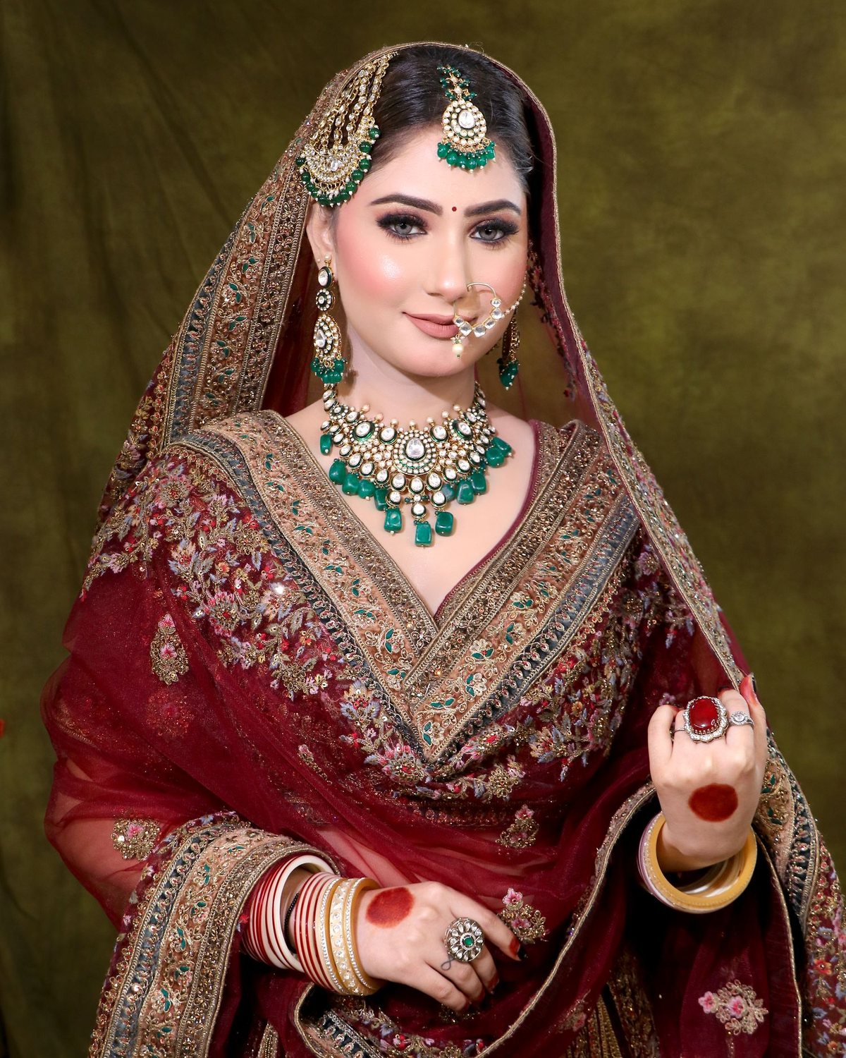 South Asian bride in a deep maroon lehenga with gold and emerald jewellery, wearing soft glam bridal makeup